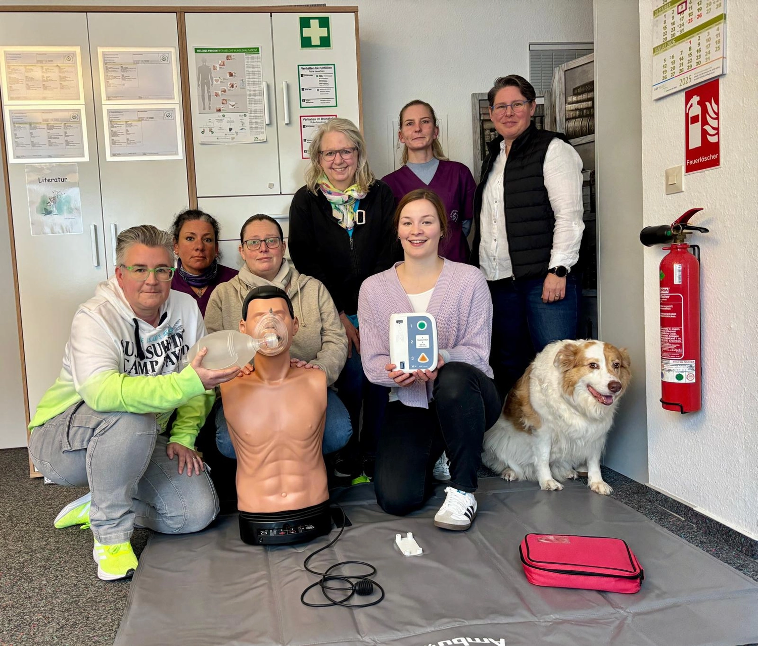 Pflegedienst Döring - Ambulanten Pflegedienstes - Eine Gruppe von Menschen posiert während einer Erste-Hilfe-Schulung für ein Foto, darunter eine Reanimationspuppe, medizinische Ausrüstung und ein Hund.
