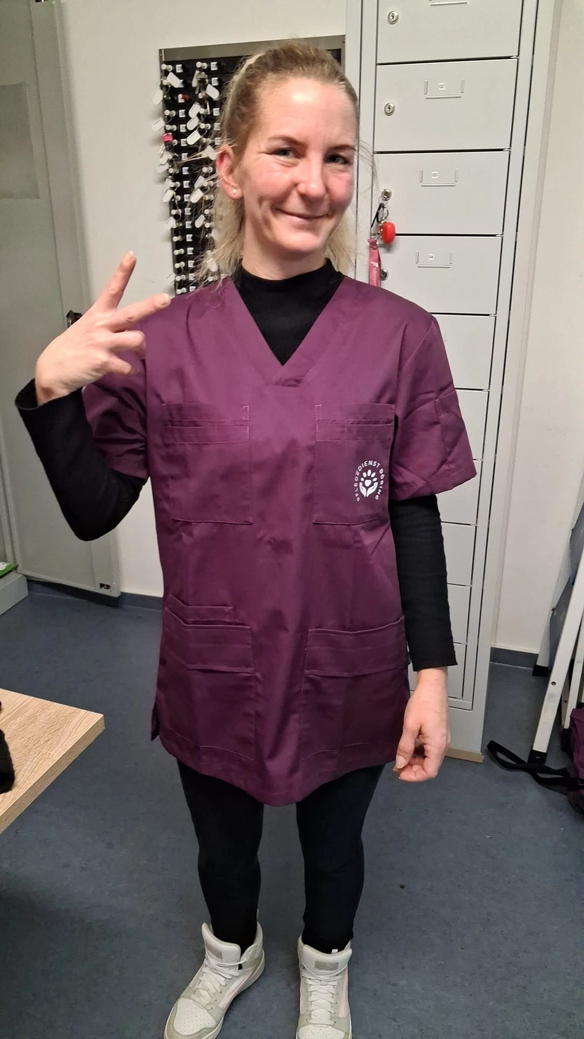 Pflegedienst Döring - Ambulanten Pflegedienstes - Eine Frau mit zurückgebundenem blonden Haar, die über einem schwarzen Langarmshirt und schwarzen Leggings eine violette Arbeitskleidung mit V-Ausschnitt trägt, lächelt und zeigt in einer Umkleidekabine das Friedenszeichen.