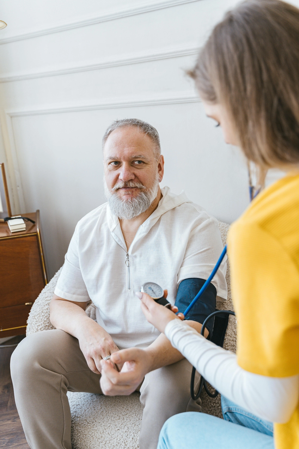 Pflegedienst Döring - Ambulanten Pflegedienstes - Eine weibliche Gesundheitshelferin in einem gelben Hemd misst den Blutdruck eines Mannes mittleren Alters mit grauem Bart mit einem manuellen Blutdruckmessgerät.