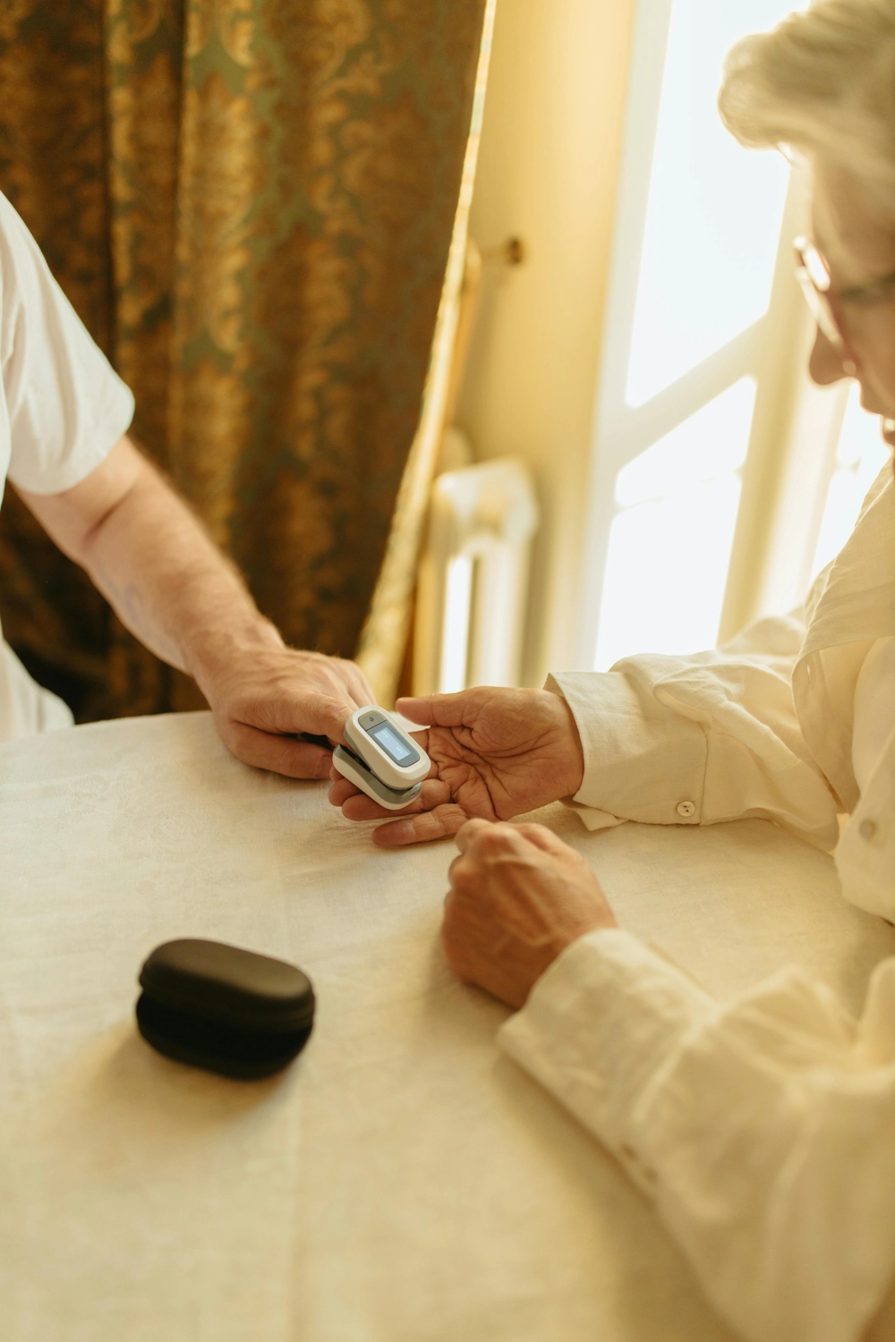 Pflegedienst Döring - Ambulanten Pflegedienstes - Eine ältere Frau sitzt an einem sonnenbeschienenen Tisch, während eine Pflegekraft oder ein Verwandter ihren Gesundheitszustand mit einem Pulsoximeter an ihrem Finger überwacht.
