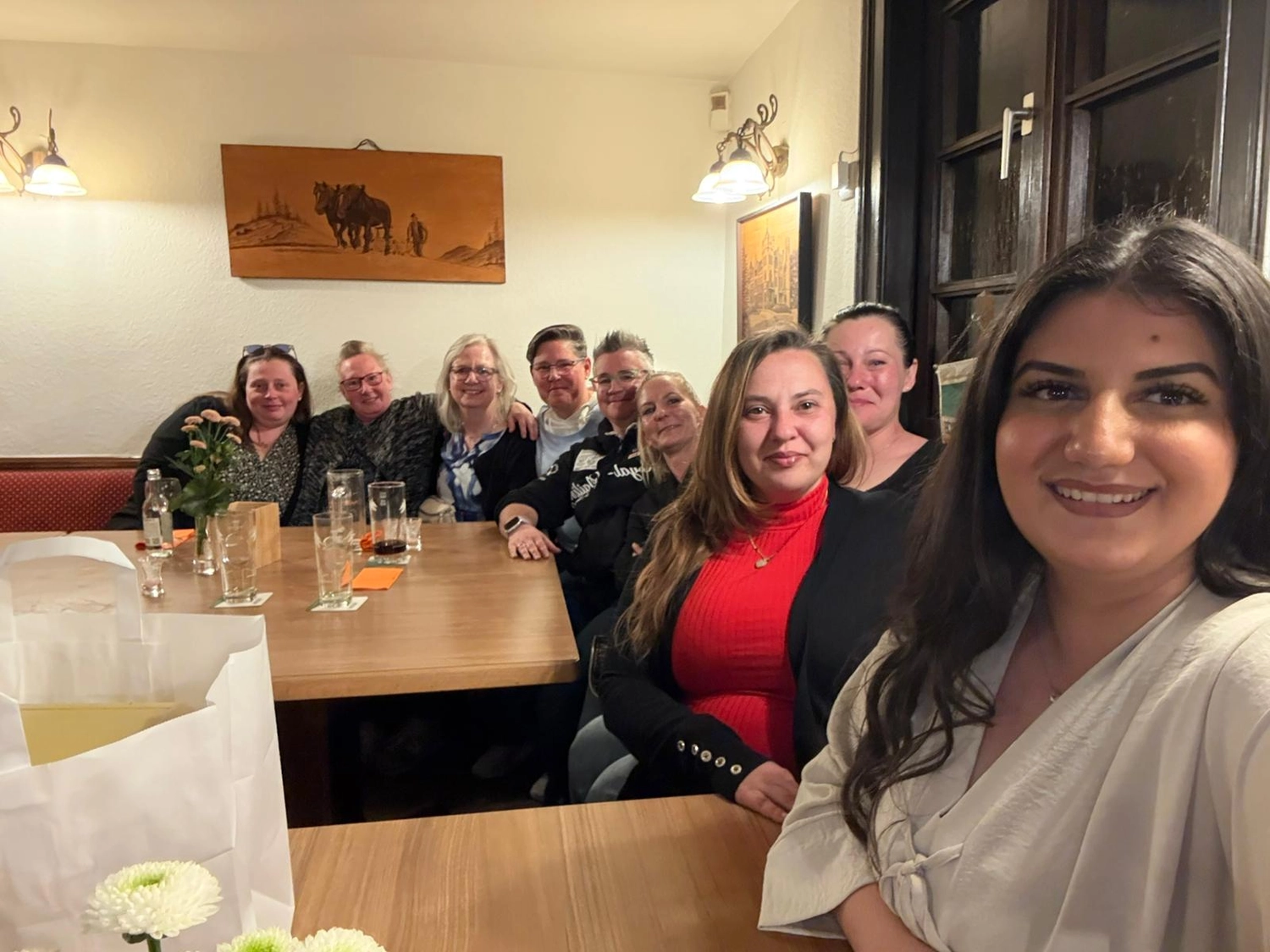 Pflegedienst Döring - Ambulanten Pflegedienstes - In einem hell erleuchteten Restaurant mit weißen Wänden und dunklen Holzelementen sitzen Frauen um einen großen Holztisch. Die Tische sind mit Gläsern und kleinen Blumenarrangements gedeckt, während gerahmte Kunstwerke und eine Wandlampe den Hintergrund schmücken.