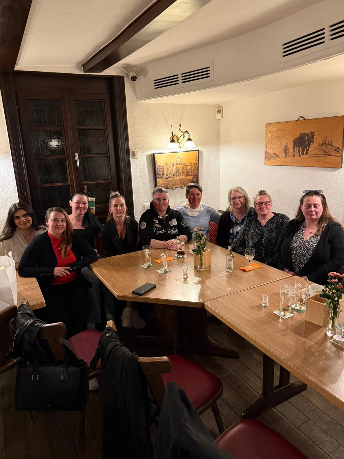 Pflegedienst Döring - Ambulanten Pflegedienstes - In einem hell erleuchteten Restaurant mit weißen Wänden und dunklen Holzelementen sitzen Frauen um einen großen Holztisch. Die Tische sind mit Gläsern und kleinen Blumenarrangements gedeckt, während gerahmte Kunstwerke und eine Wandlampe den Hintergrund schmücken.